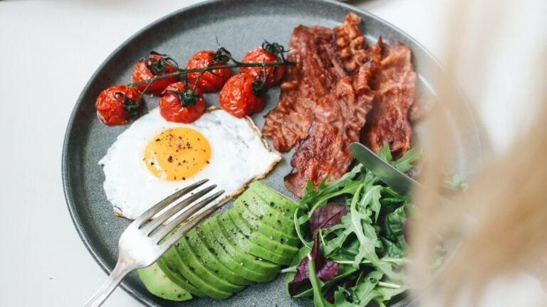 A charcoal coloured plate on a white table. There is a fried egg, a sliced avocado, about 4 slices of bacon, a few cherry tomatoes and some salad greens on the plate.