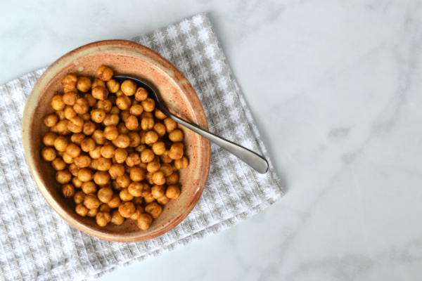 a bowl filled with roasted chickpeas and a small spoon sitting on top of a dish towel and a marble countertop