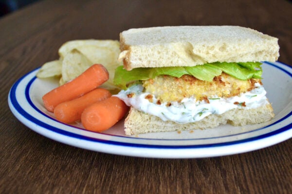 a breaded fish sandwich sitting on a white and blue plate with carrot sticks and some plain potato chips