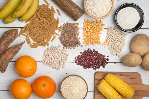 several starch foods are scattered on a wooden table including bananas, pasta, oranges, corn, potatoes, and flour