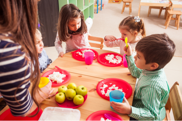 four children sitting at a table eating lunch with an adult helping to feed them